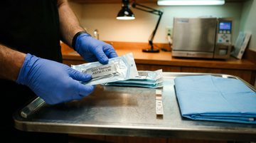 Piercer's gloved hands holding a sealed sterile G23 implant-grade titanium piercing jewelry pouch on a stainless steel tray with autoclave indicator strip — SSZ Piercing material safety guide