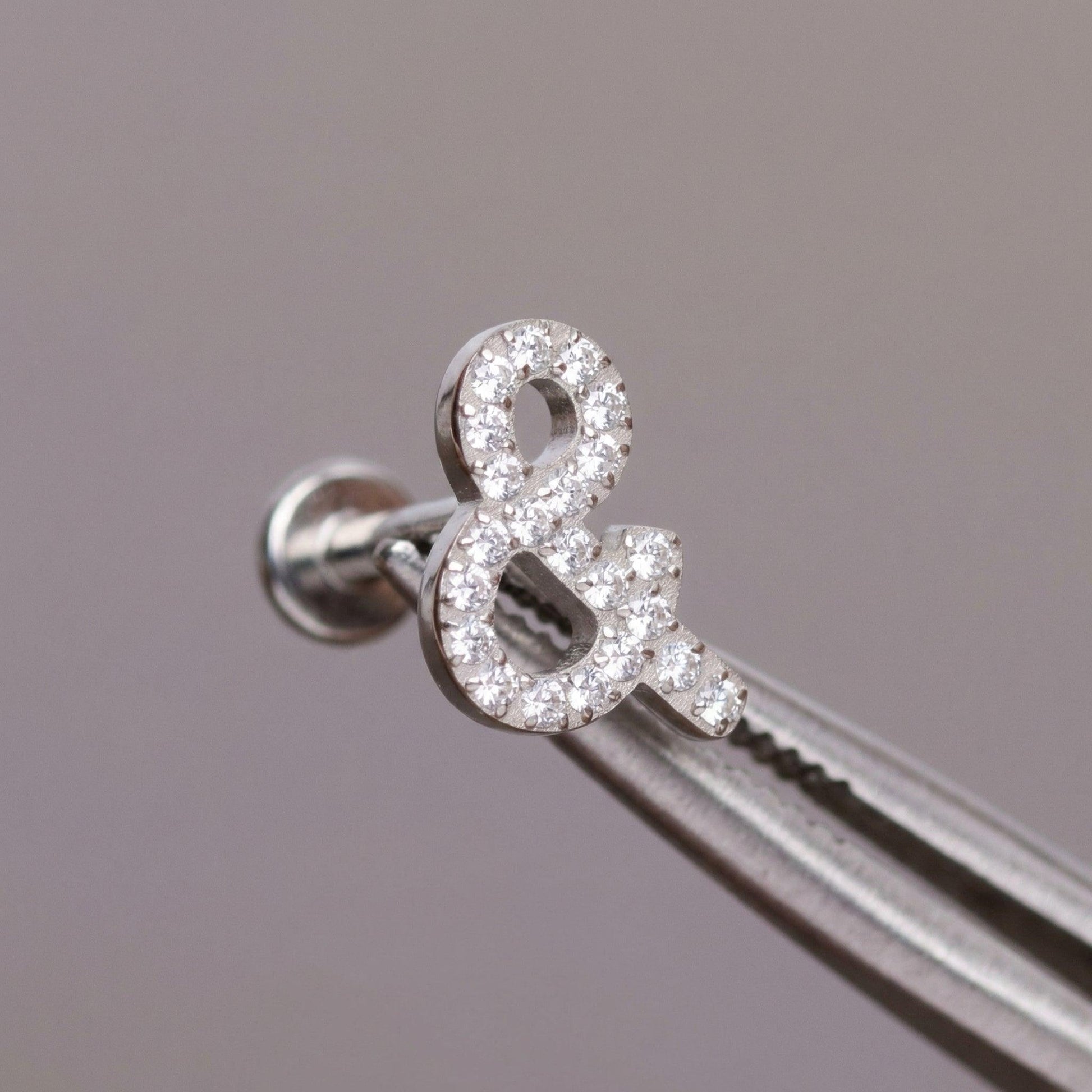 Tweezers holding a silver titanium ampersand cartilage piercing stud.