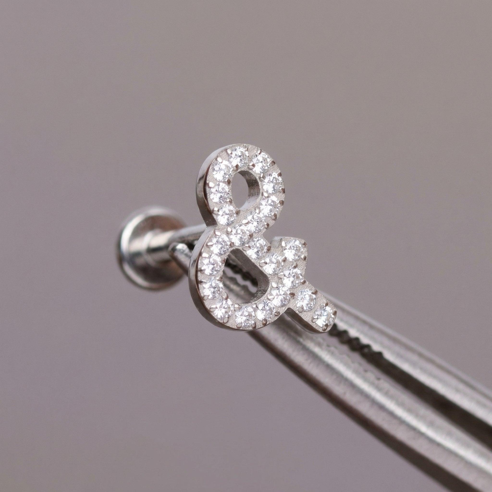 Tweezers holding a silver titanium ampersand cartilage piercing stud.