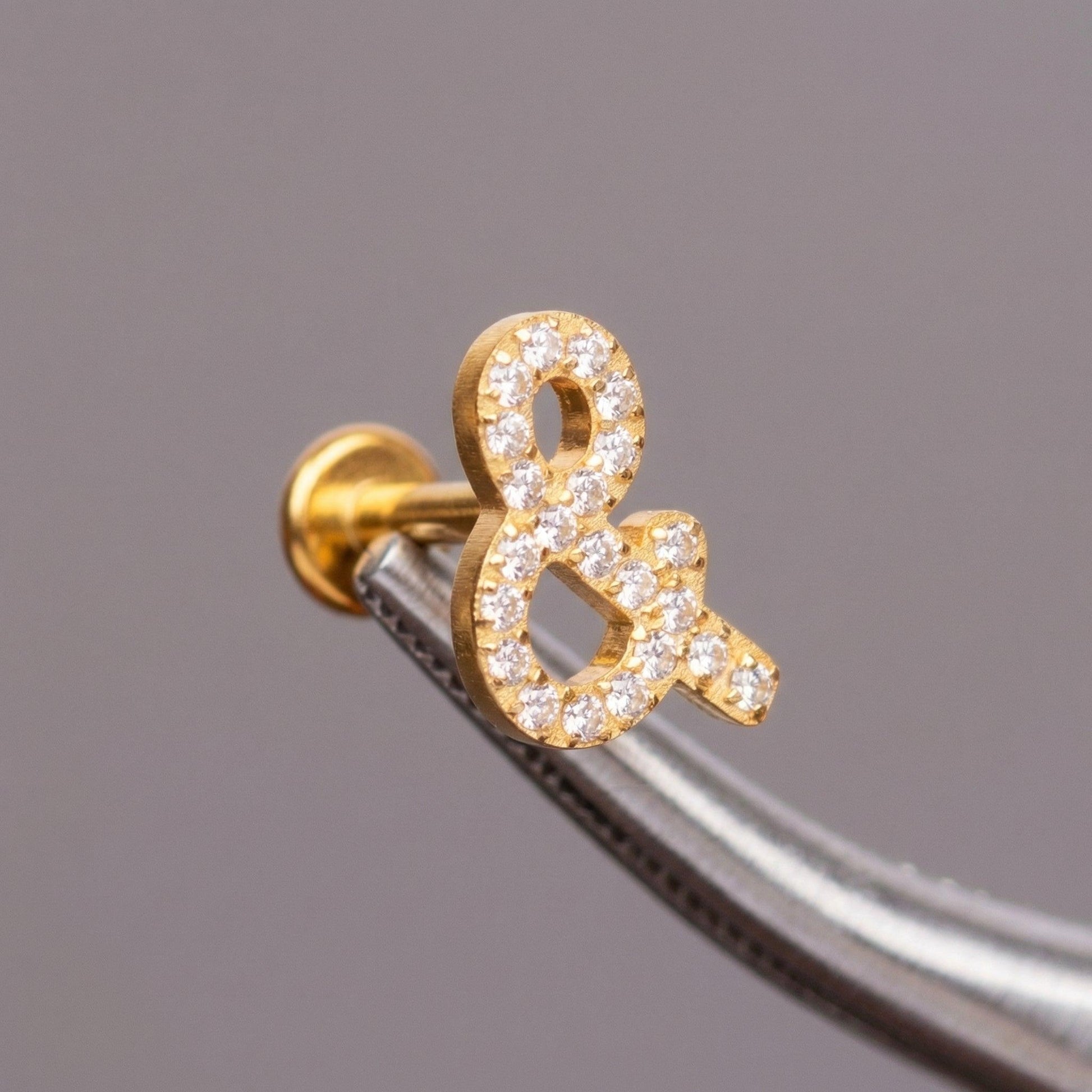 Tweezers holding a gold titanium ampersand cartilage piercing stud.