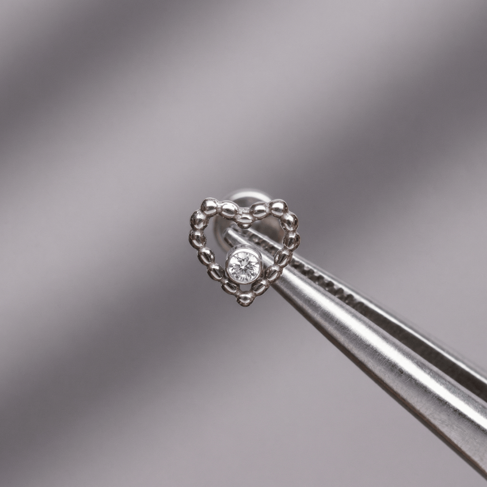 Tweezers holding a silver titanium beaded heart cartilage piercing stud against a