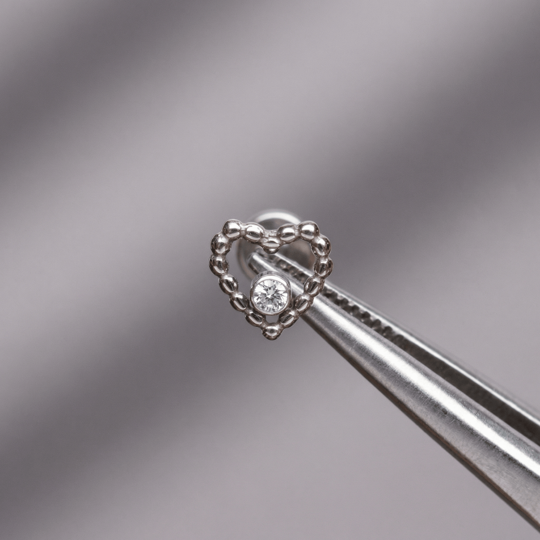 Tweezers holding a silver titanium beaded heart cartilage piercing stud against a