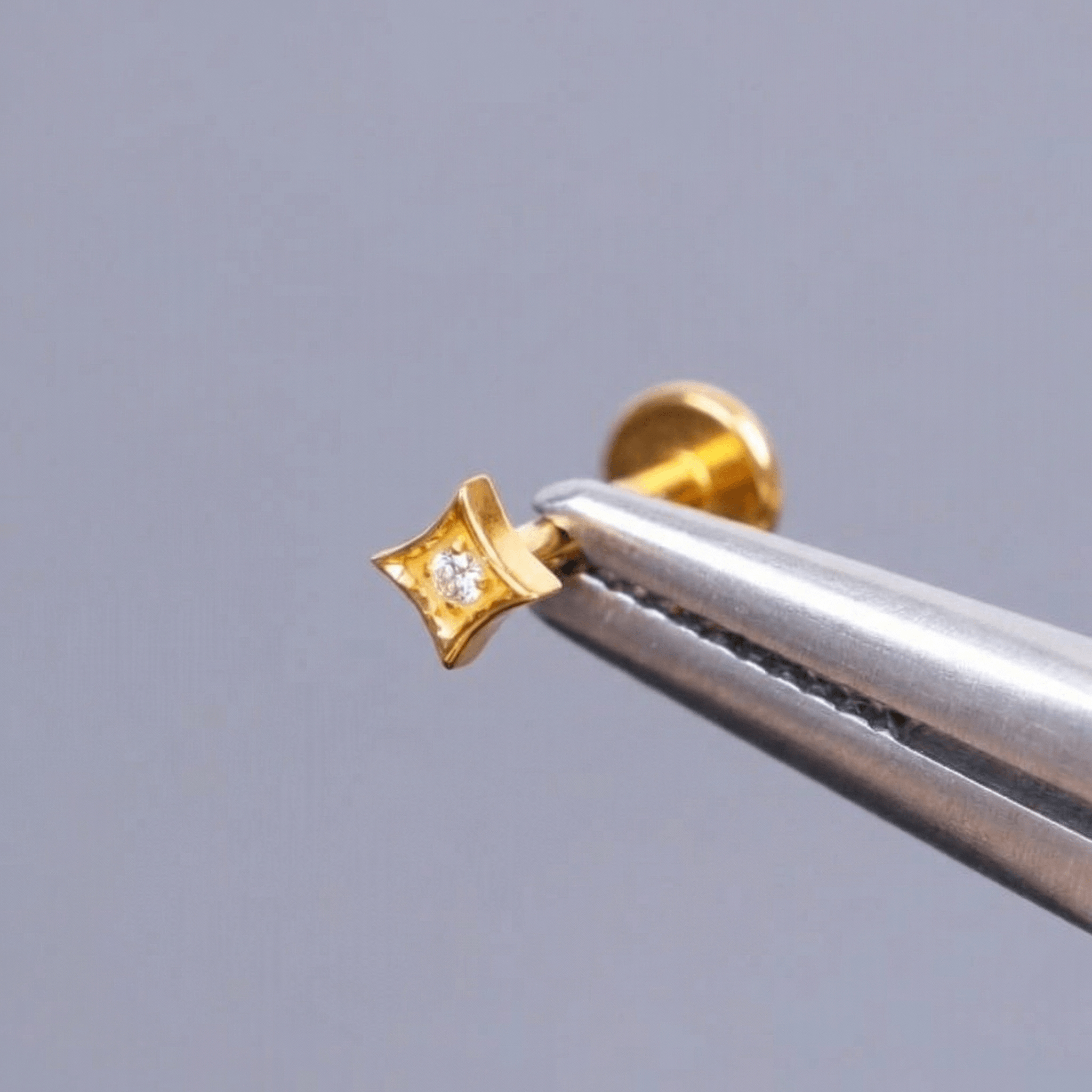 Gold geometric piercing stud held securely by metal tweezers.
