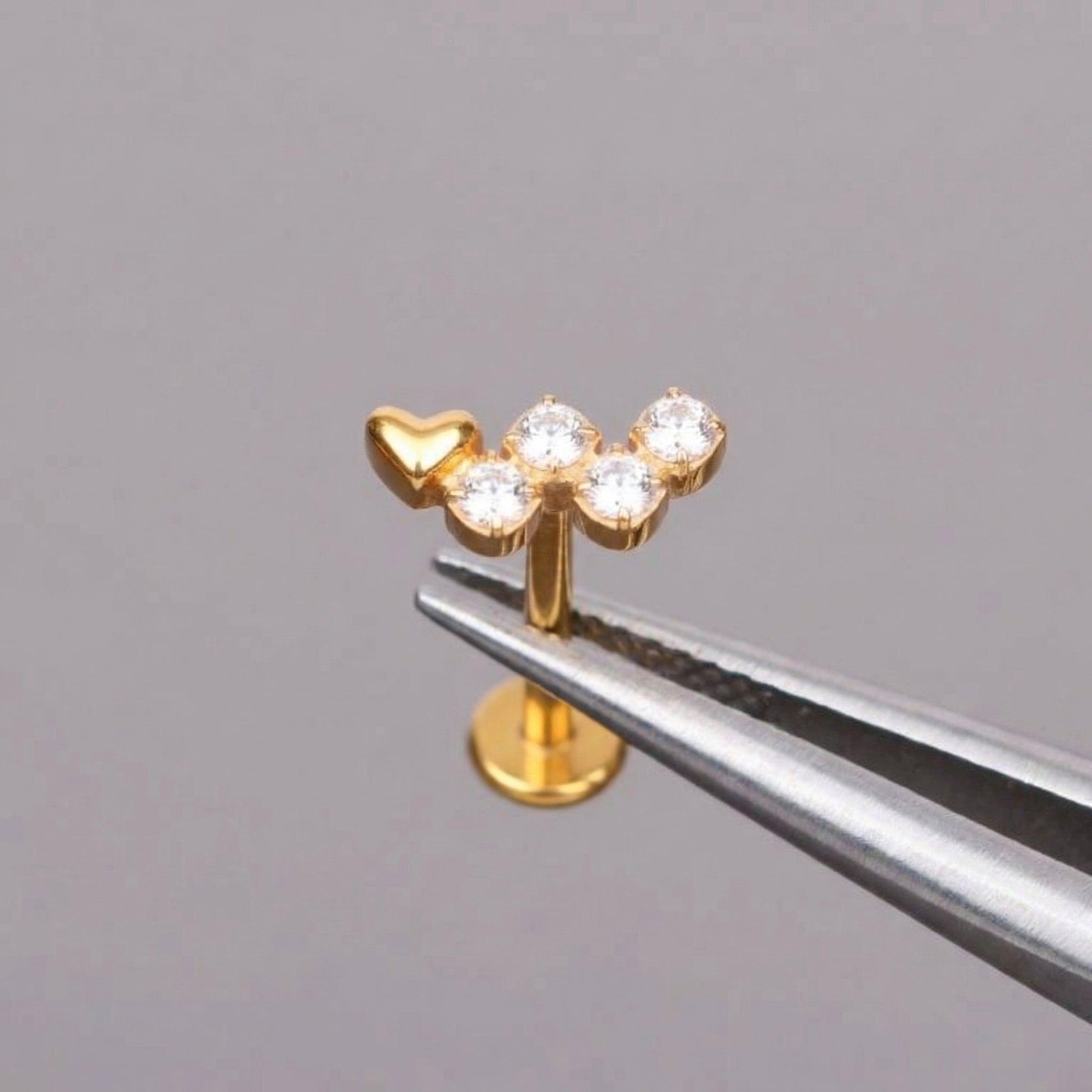 Gold heart clear stone cluster stud held safely with metal piercing tweezers.