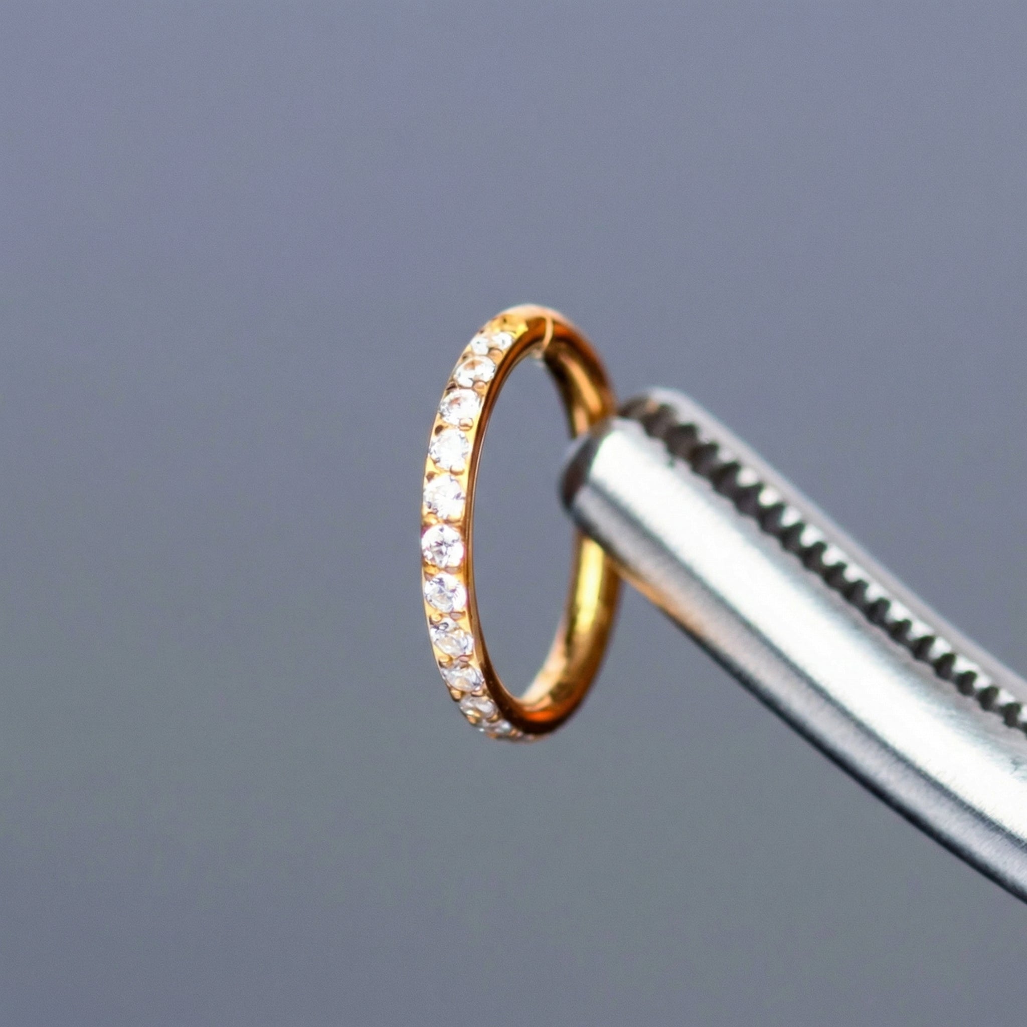 Tweezers holding a gold titanium zircon clicker hoop ring against a gray