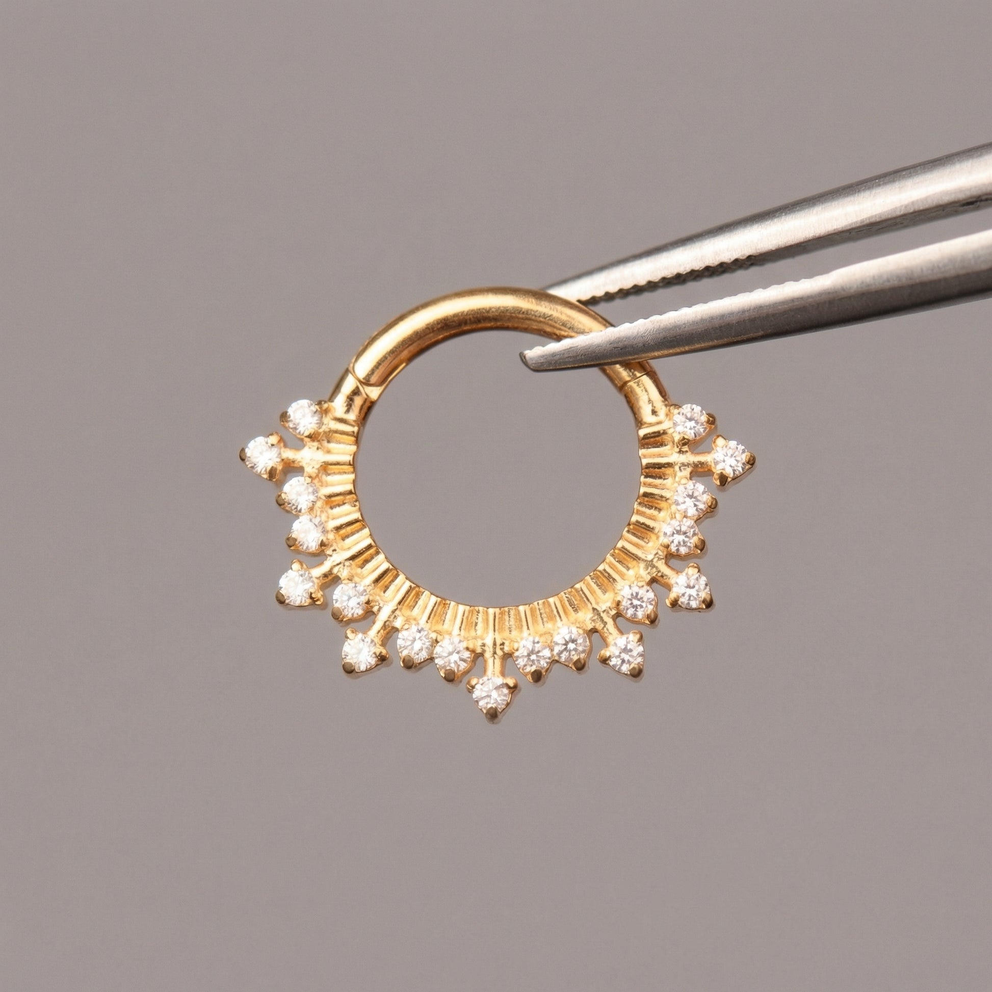 Precision tweezers holding a gold titanium zircon crown hinged clicker hoop ring.