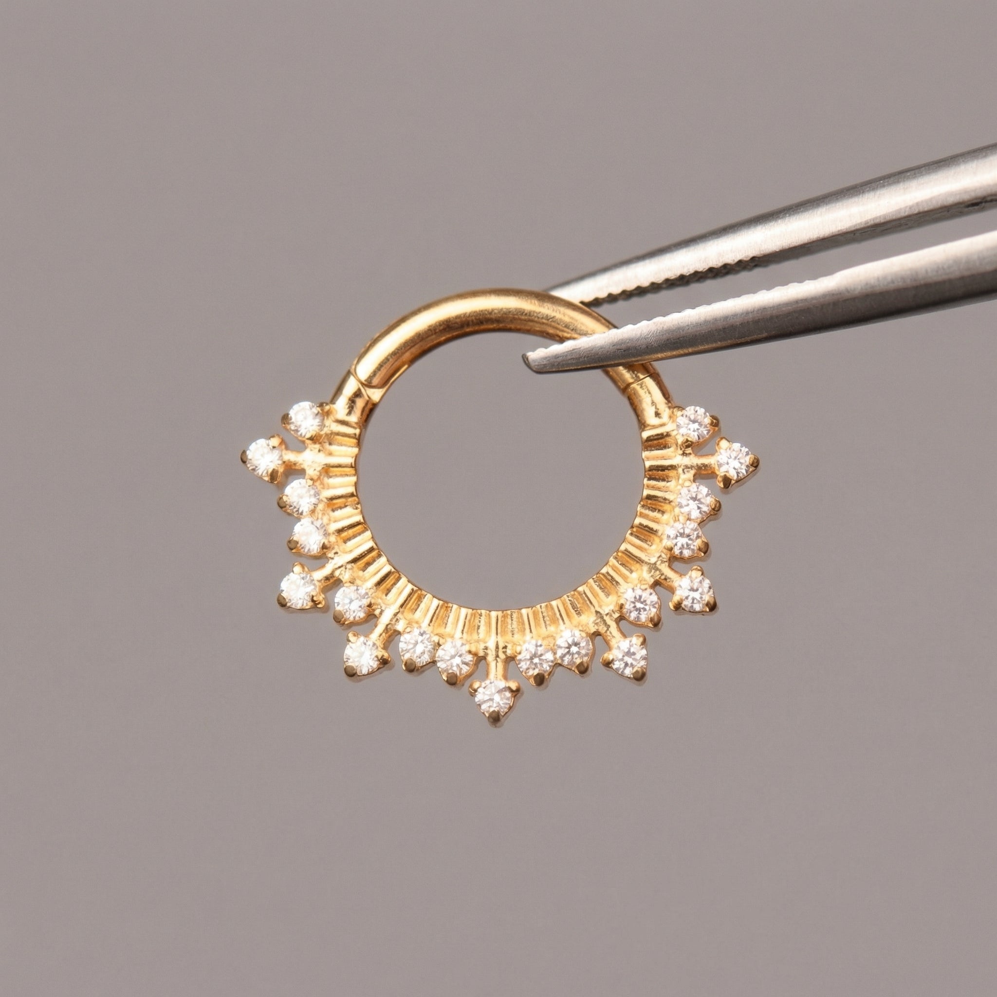 Precision tweezers holding a gold titanium zircon crown hinged clicker hoop ring.